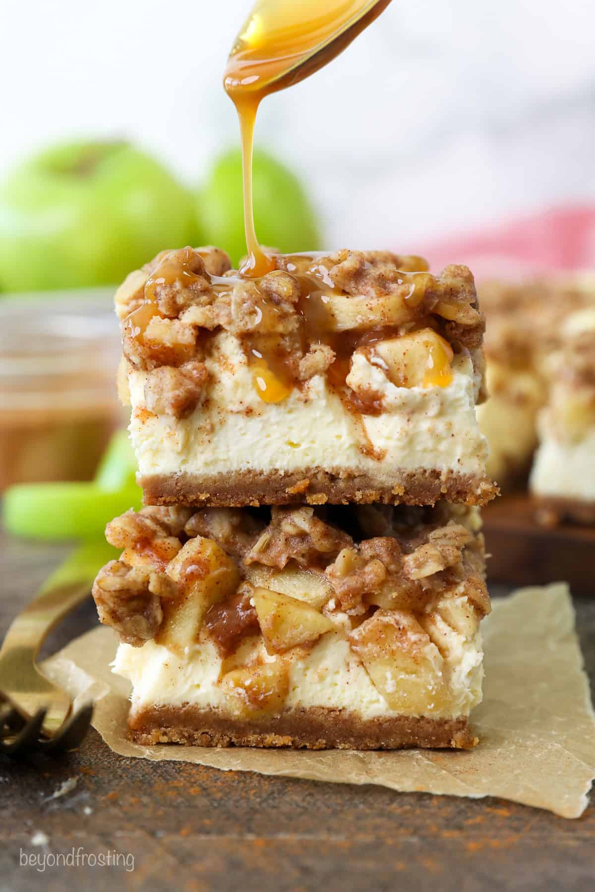 Two caramel apple cheesecake bars stacked on top of one another, being drizzled with caramel sauce.