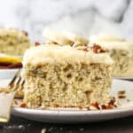 A slice of frosted banana cake topped with fresh banana slices on a plate next to a fork, with more slices in the background.