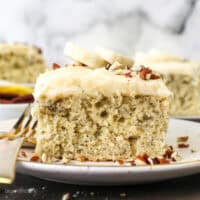 A slice of frosted banana cake topped with fresh banana slices on a plate next to a fork, with more slices in the background.