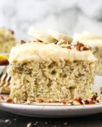 A slice of frosted banana cake topped with fresh banana slices on a plate next to a fork, with more slices in the background.