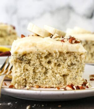 A slice of frosted banana cake topped with fresh banana slices on a plate next to a fork, with more slices in the background.