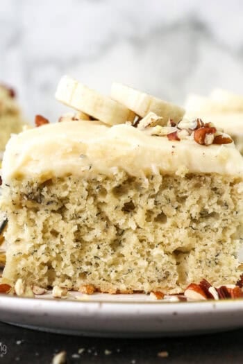 A slice of frosted banana cake topped with fresh banana slices on a plate next to a fork, with more slices in the background.