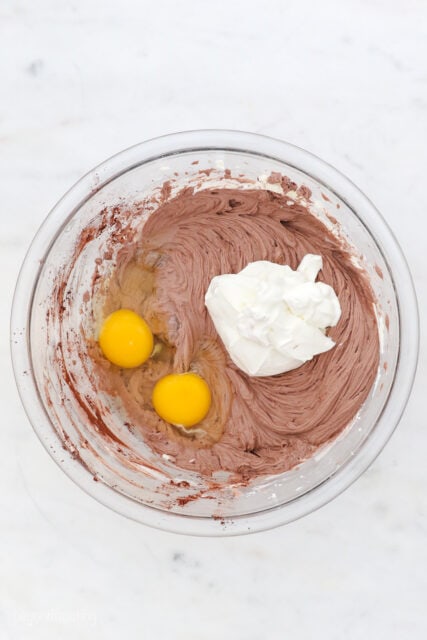 Eggs and whipped cream added to chocolate cheesecake batter in a glass mixing bowl.