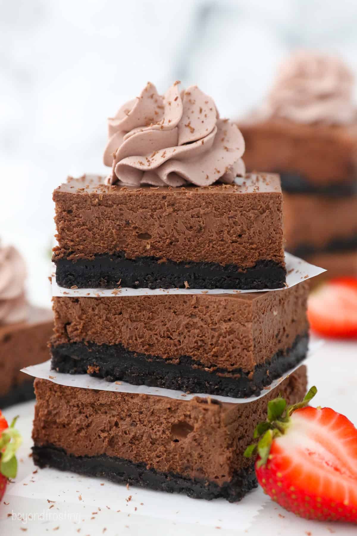 A stack of three chocolate cheesecake bars topped with a swirl of chocolate whipped cream, next to a halved strawberry.