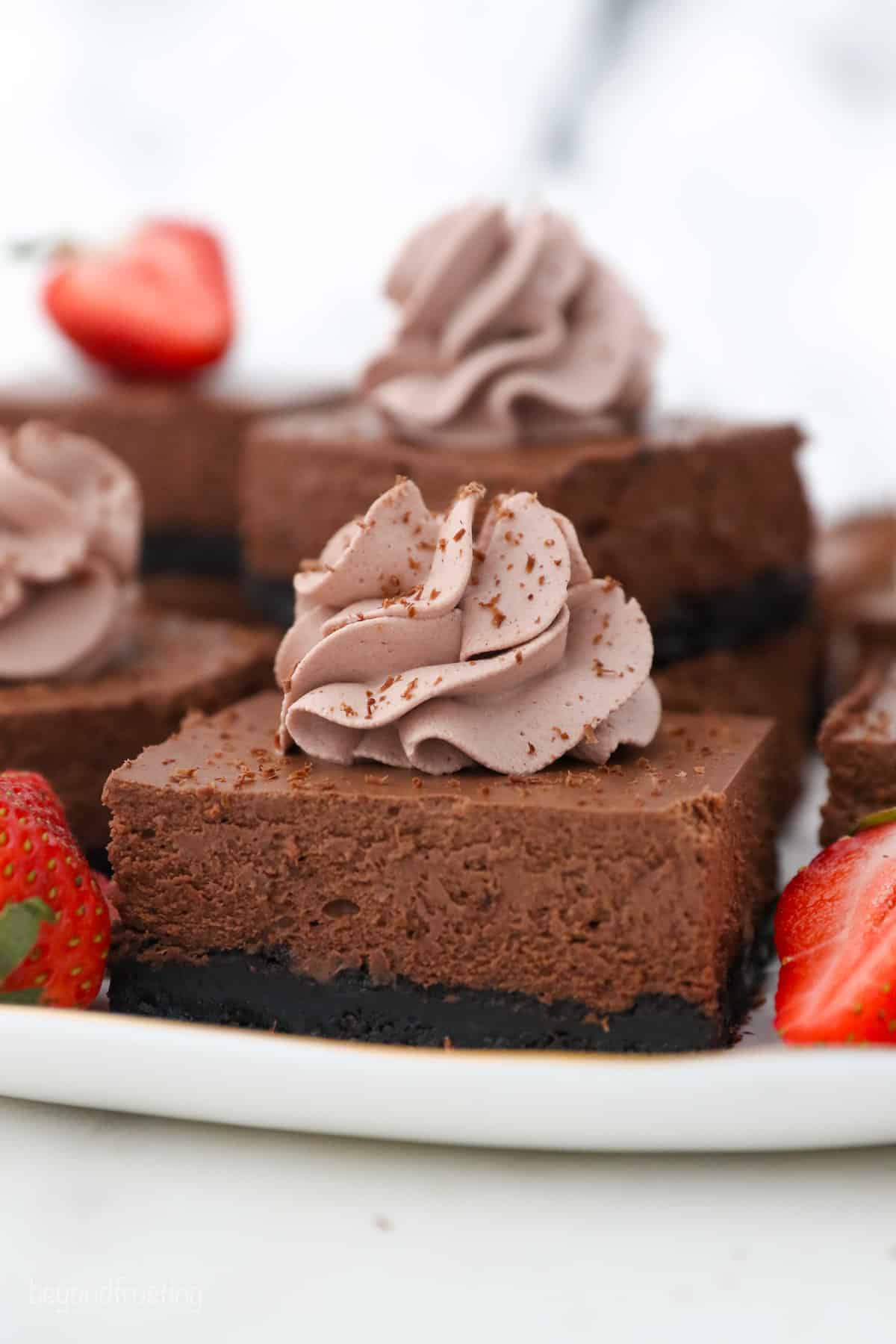 Close up of chocolate cheesecake bars topped with swirls of chocolate whipped cream, next to strawberry halves on a white platter.