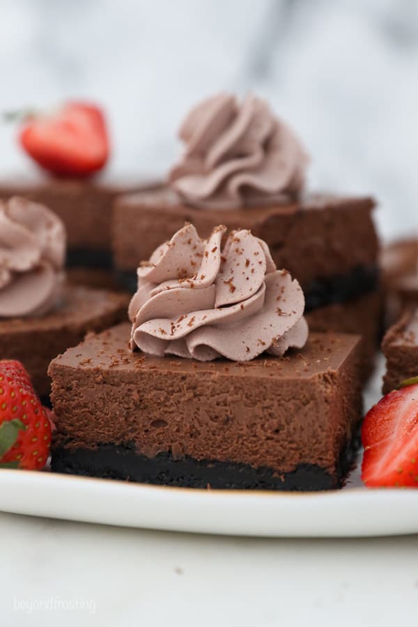 a serving plate with chocolate cheesecake bars topped with chocolate whipepd cream
