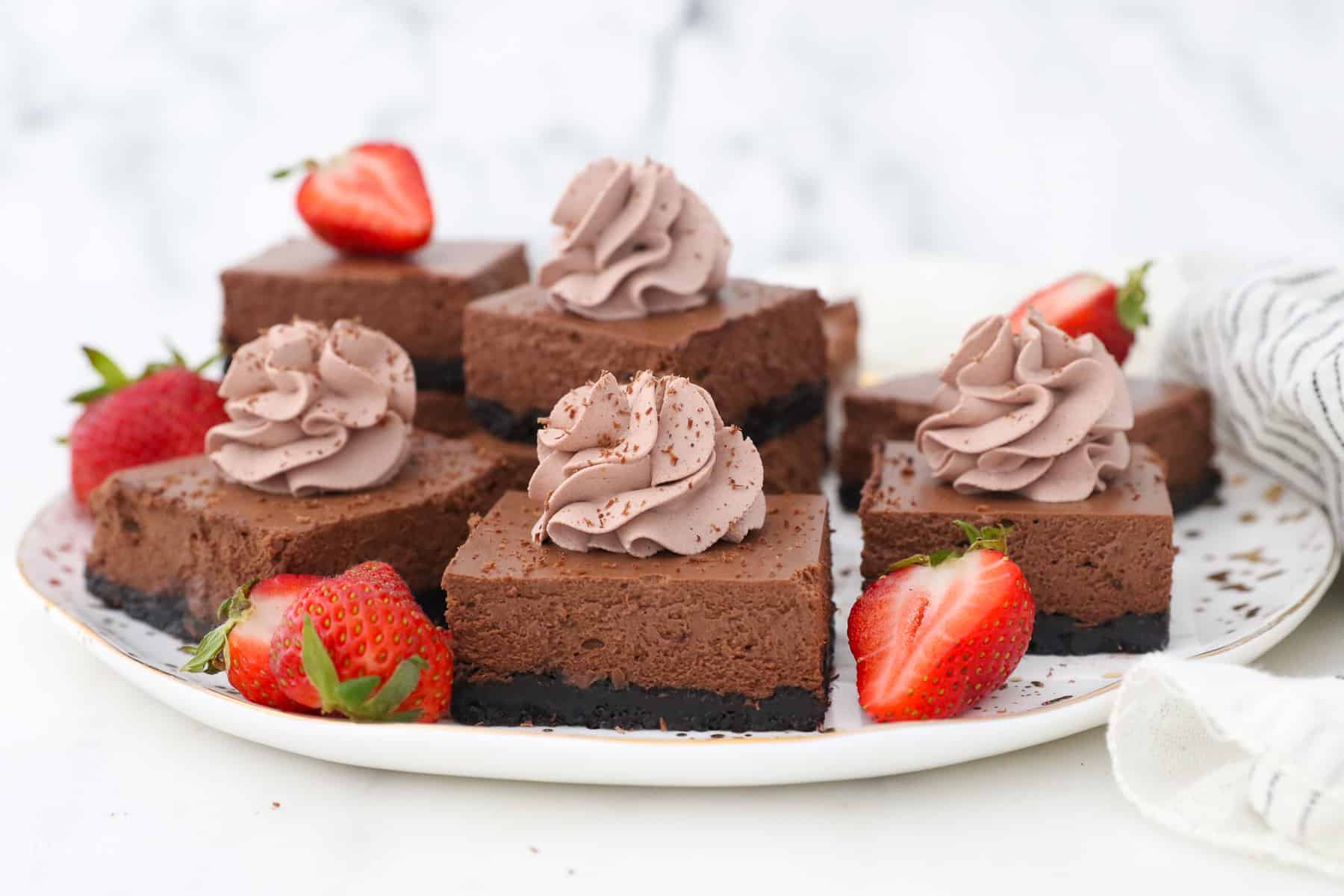 Assorted chocolate cheesecake bars topped with swirls of chocolate whipped cream, next to strawberry halves on a white platter.