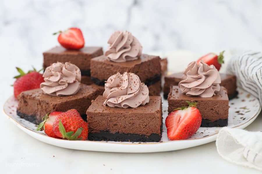 a serving plate with chocolate cheesecake bars topped with chocolate whipepd cream and strawberries