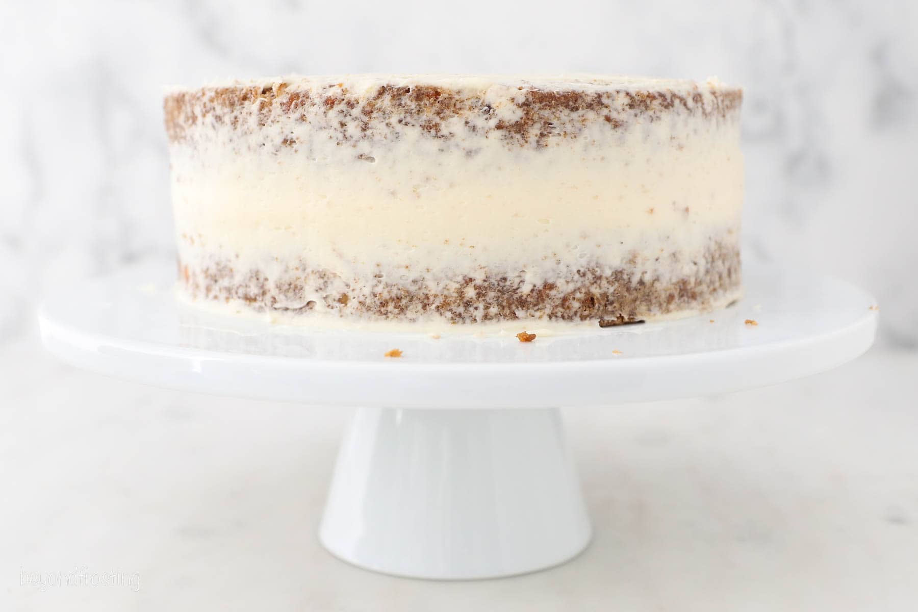 Assembled sweet potato cake with a frosting crumb coat on a white cake stand.