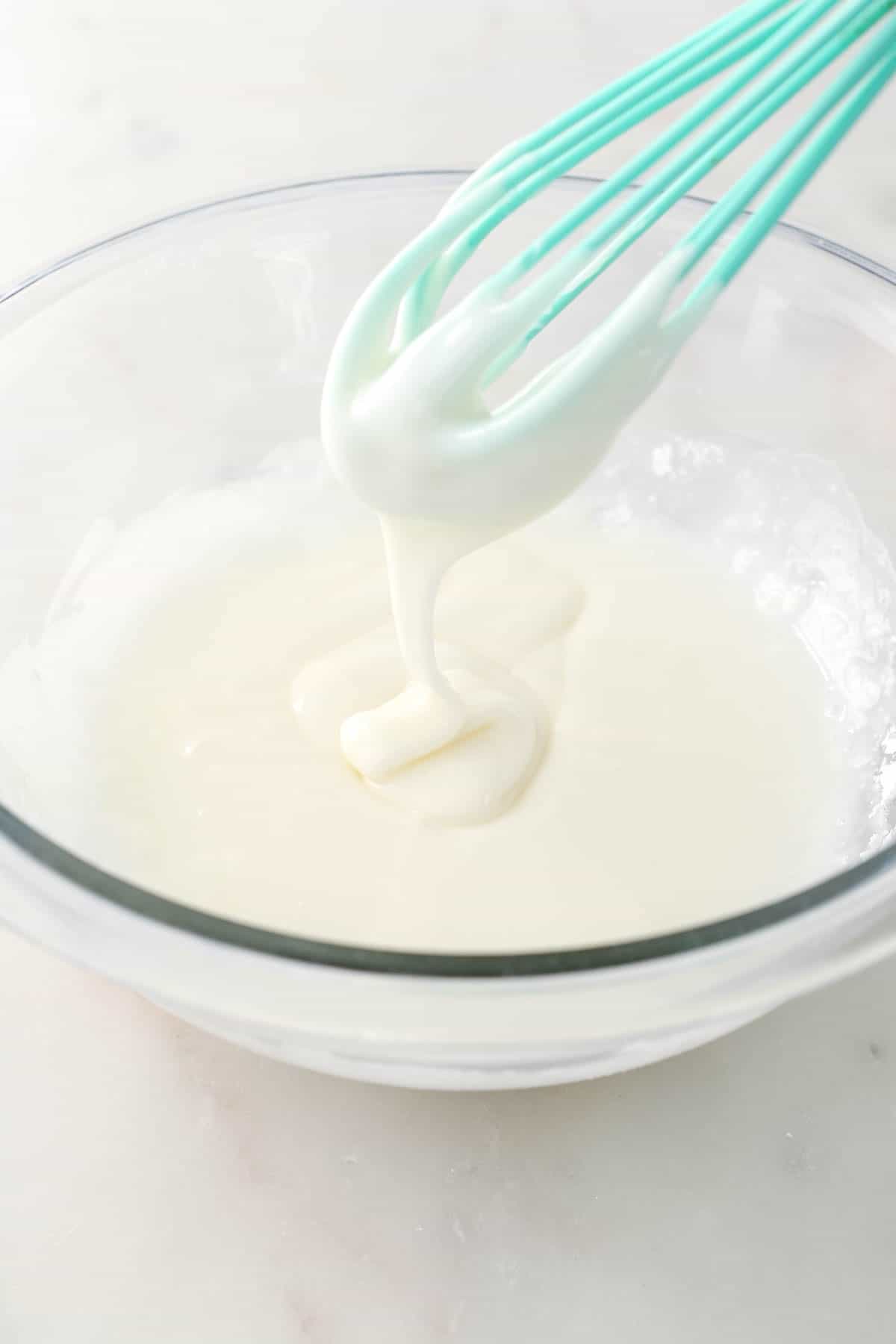 Sugar cookie icing in a glass mixing bowl, dripping from a whisk.