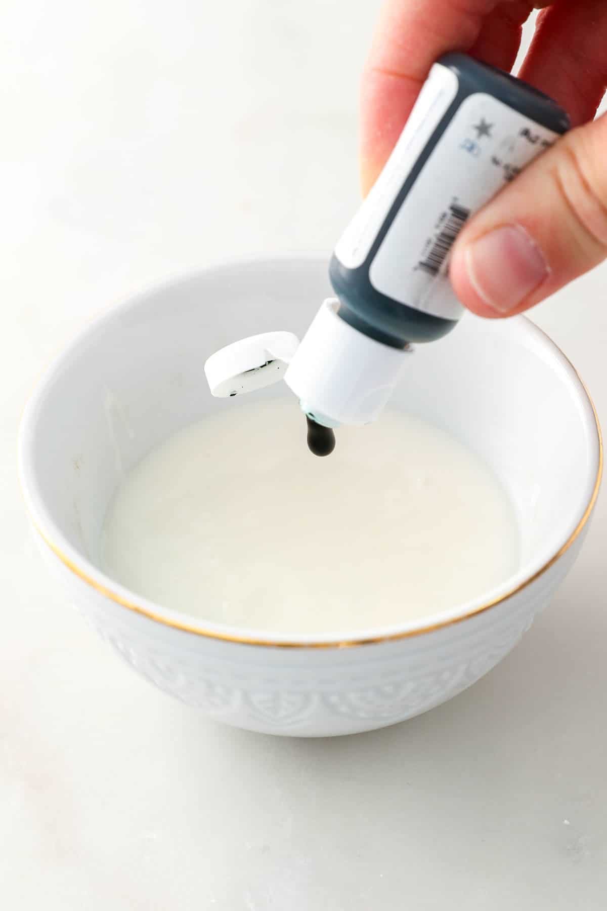 A hand squeezing a drop of green food dye into a bowl of icing.