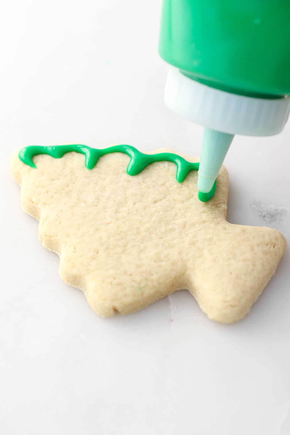 A squeeze bottle pipes green icing around the edge of a cut-out sugar cookie shaped like a Christmas tree.