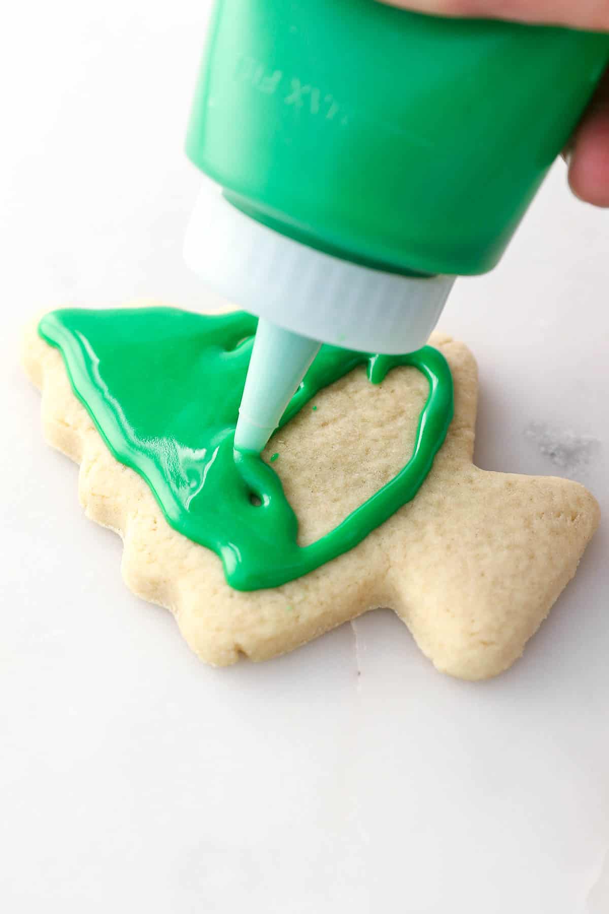 A squeeze bottle pipes green icing over a cut-out sugar cookie shaped like a Christmas tree.