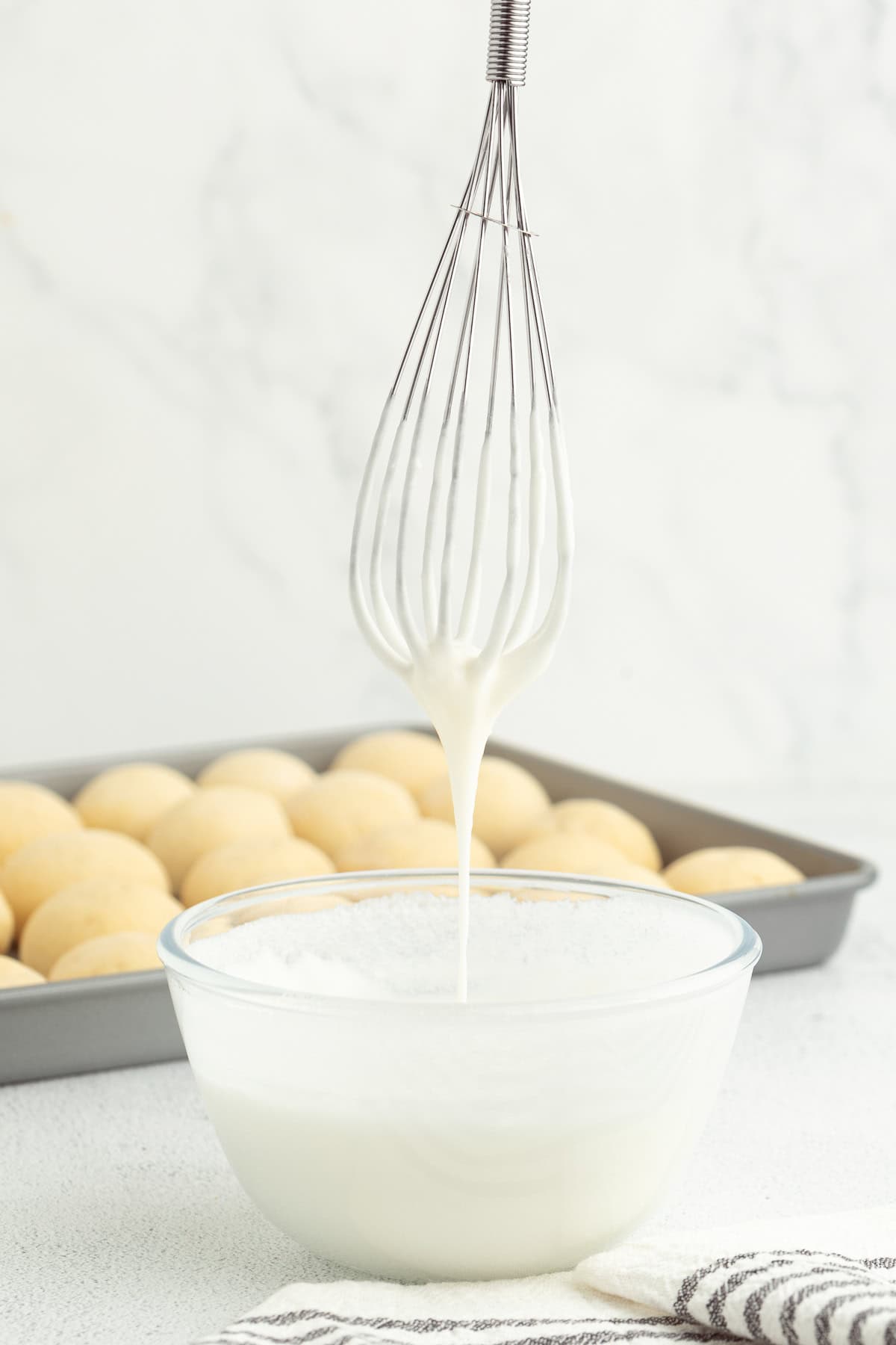 Italian anise cookies are traditional, cake-like Christmas cookies dipped in glaze and topped with sprinkles. Perfect for cookie platters! A whisk held over a bowl of icing with a tray of anise cookies in the background.