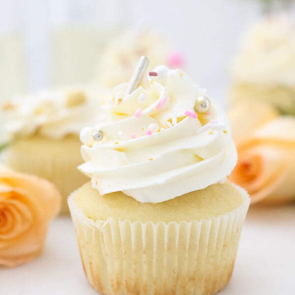 A gorgeous champagne cupcake with frosting and sprinkles and roses in the background