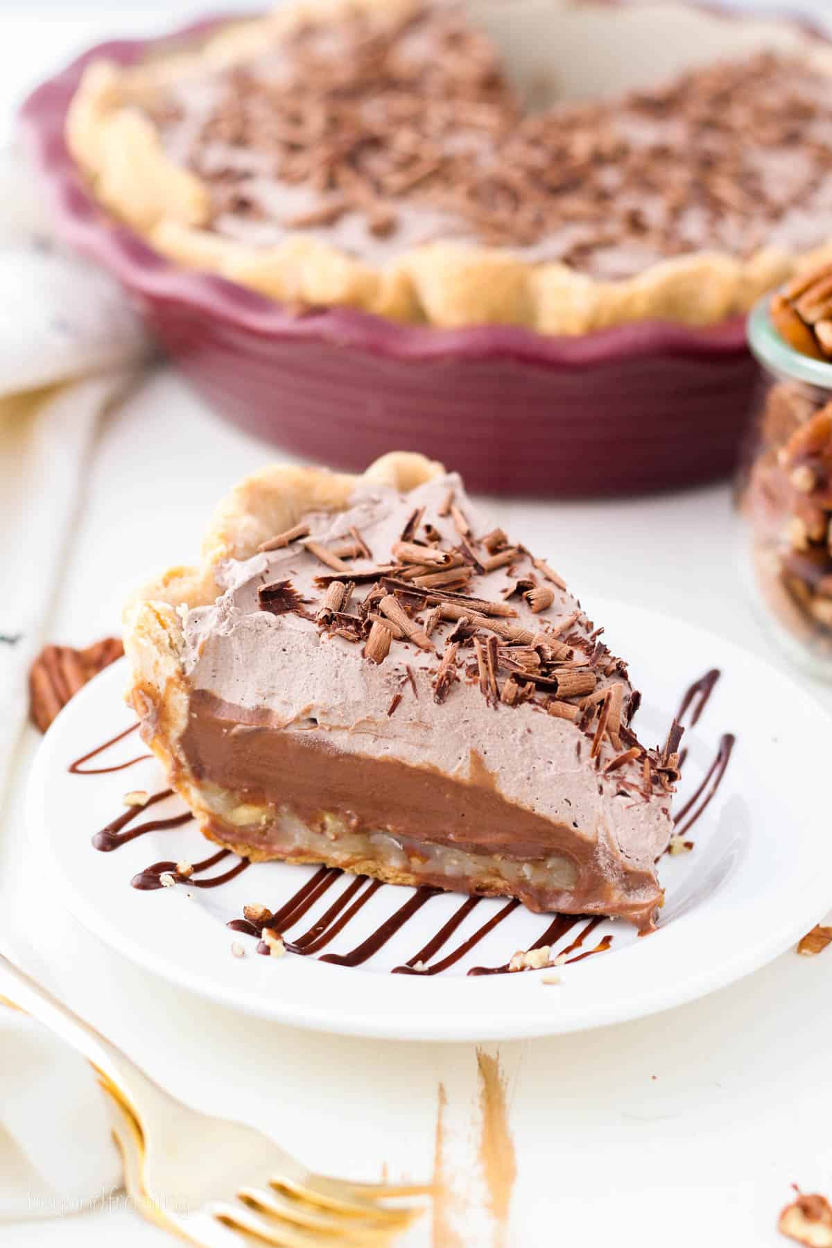A slice of German chocolate pie on a white plate drizzled with chocolate sauce, with the rest of the pie in a pie plate in the background.