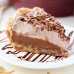 Close up of a slice of German chocolate pie on a white plate drizzled with chocolate sauce, with a pie plate in the background.