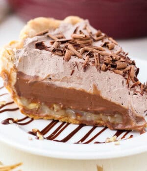 Close up of a slice of German chocolate pie on a white plate drizzled with chocolate sauce, with a pie plate in the background.