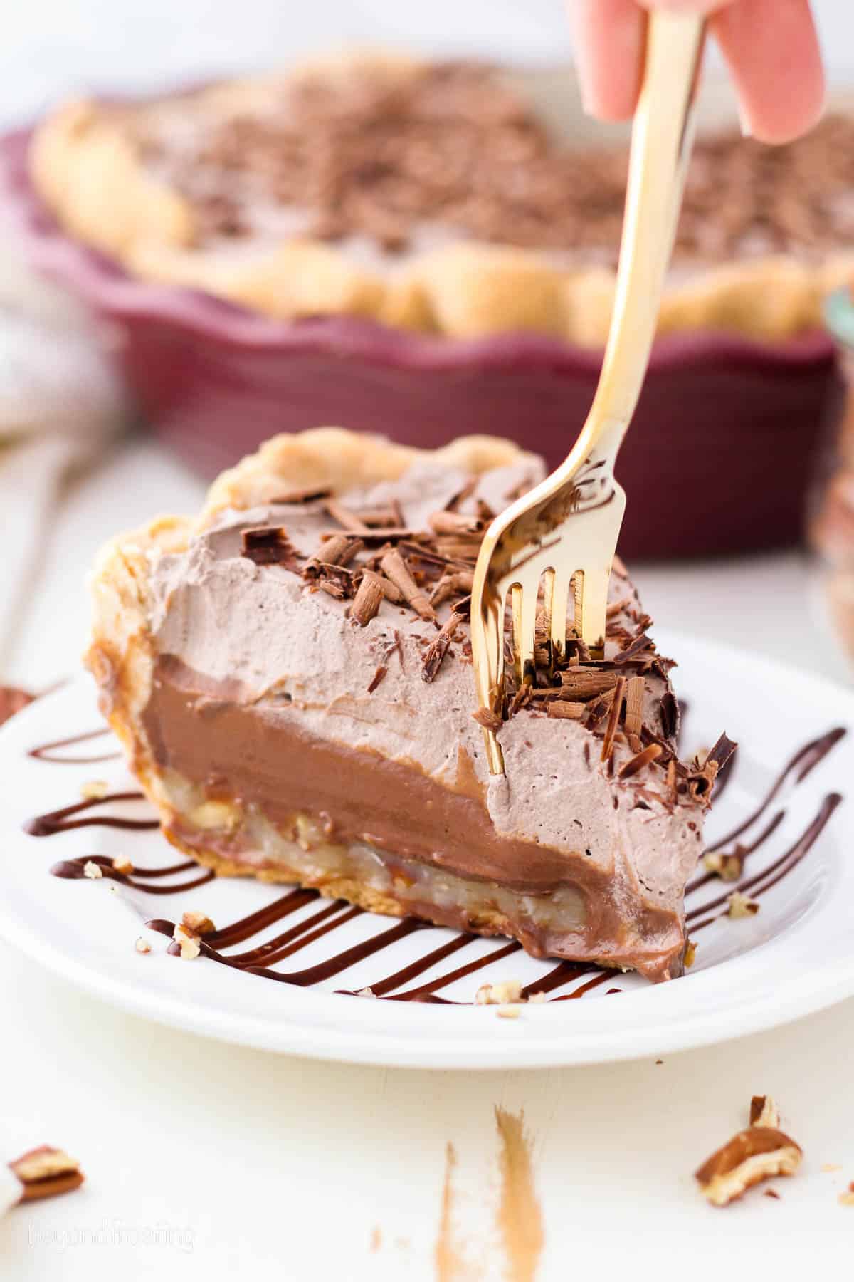 A fork stuck into the tip of a slice of German chocolate pie on a white plate drizzled with chocolate sauce, with the rest of the pie in a pie plate in the background.