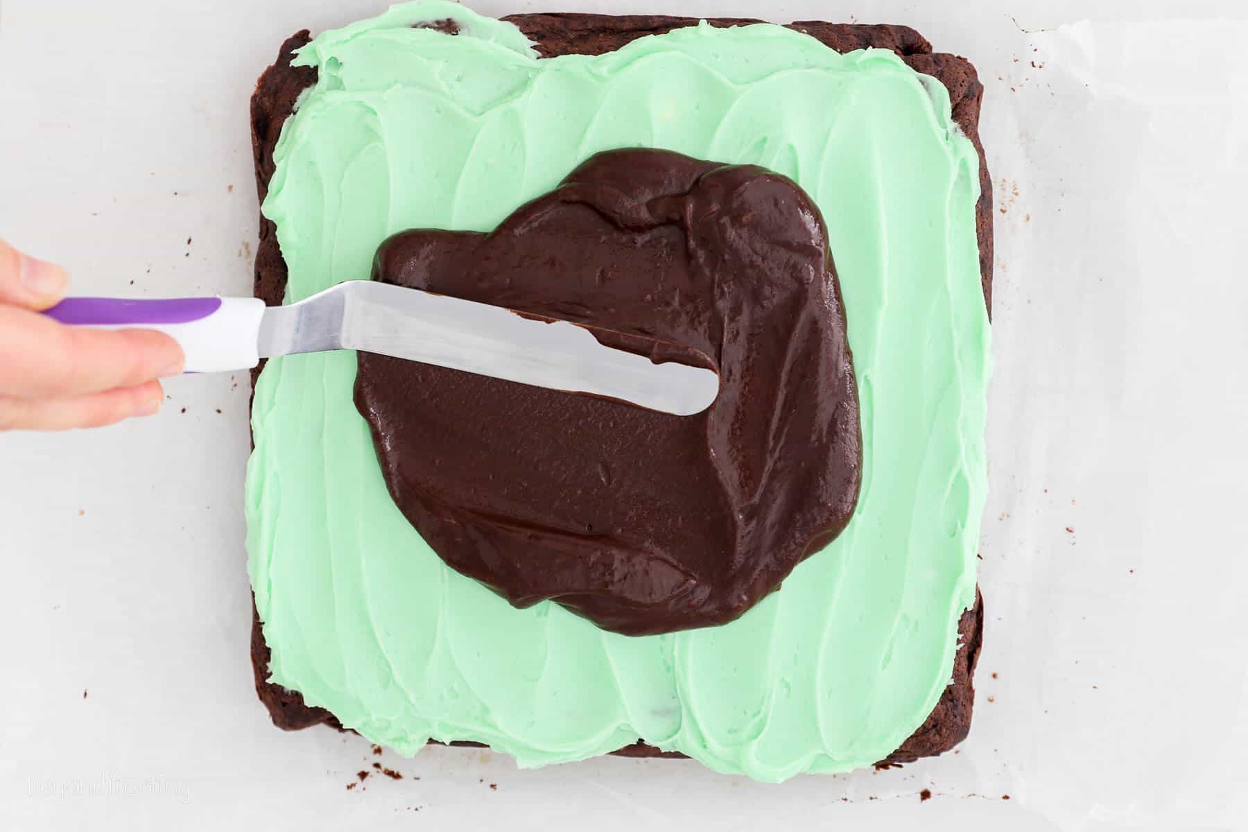 Overhead view of an offset spatula spreading chocolate ganache over top of mint chocolate brownies.