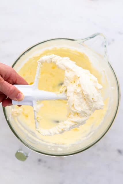 A hand holding a paddle attachment covered with whipped vanilla frosting over a bowl of frosting.