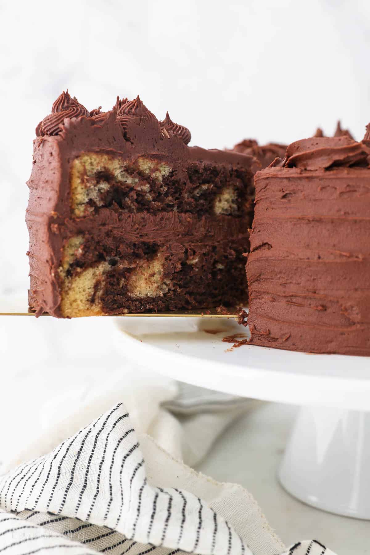 A slice of frosted marble cake is lifted from the rest of the cake on a white cake stand.