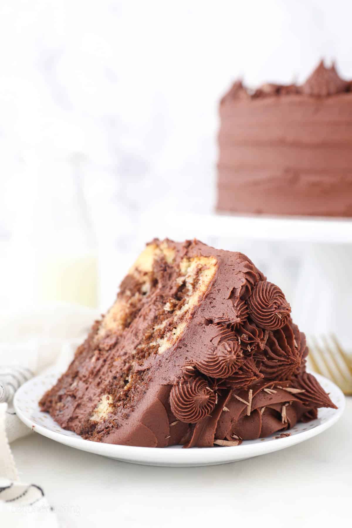 A slice of homemade frosted marble cake on a white plate, with the rest of the cake on a stand in the background.