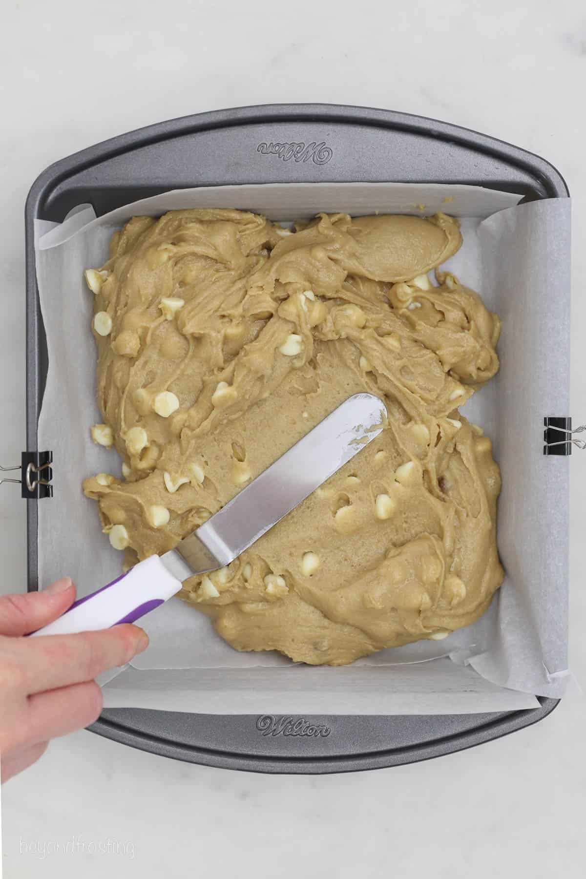 A hand uses a spatula to spread blondie batter into a square cake pan lined with parchment paper.