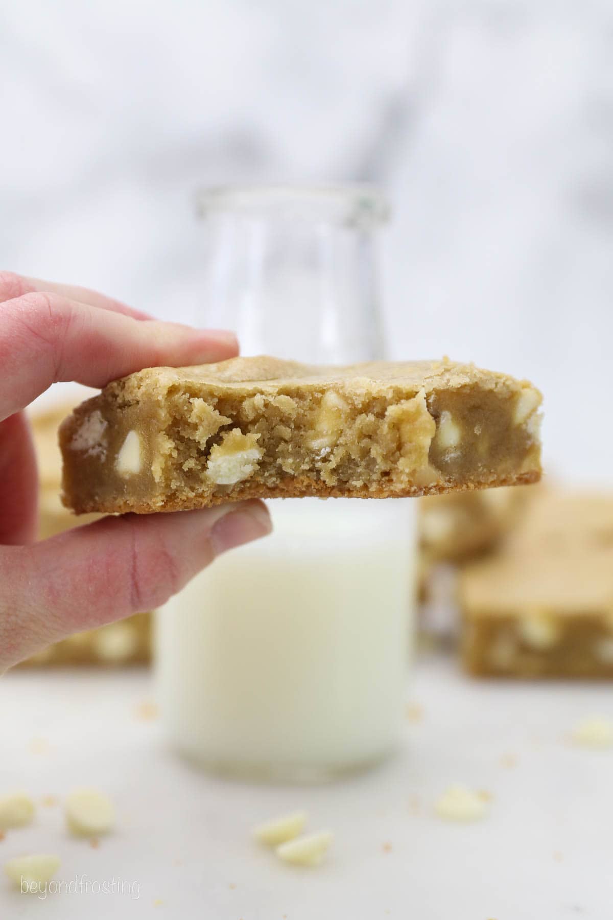 A hand holding a blondie with a glass of milk in the background.