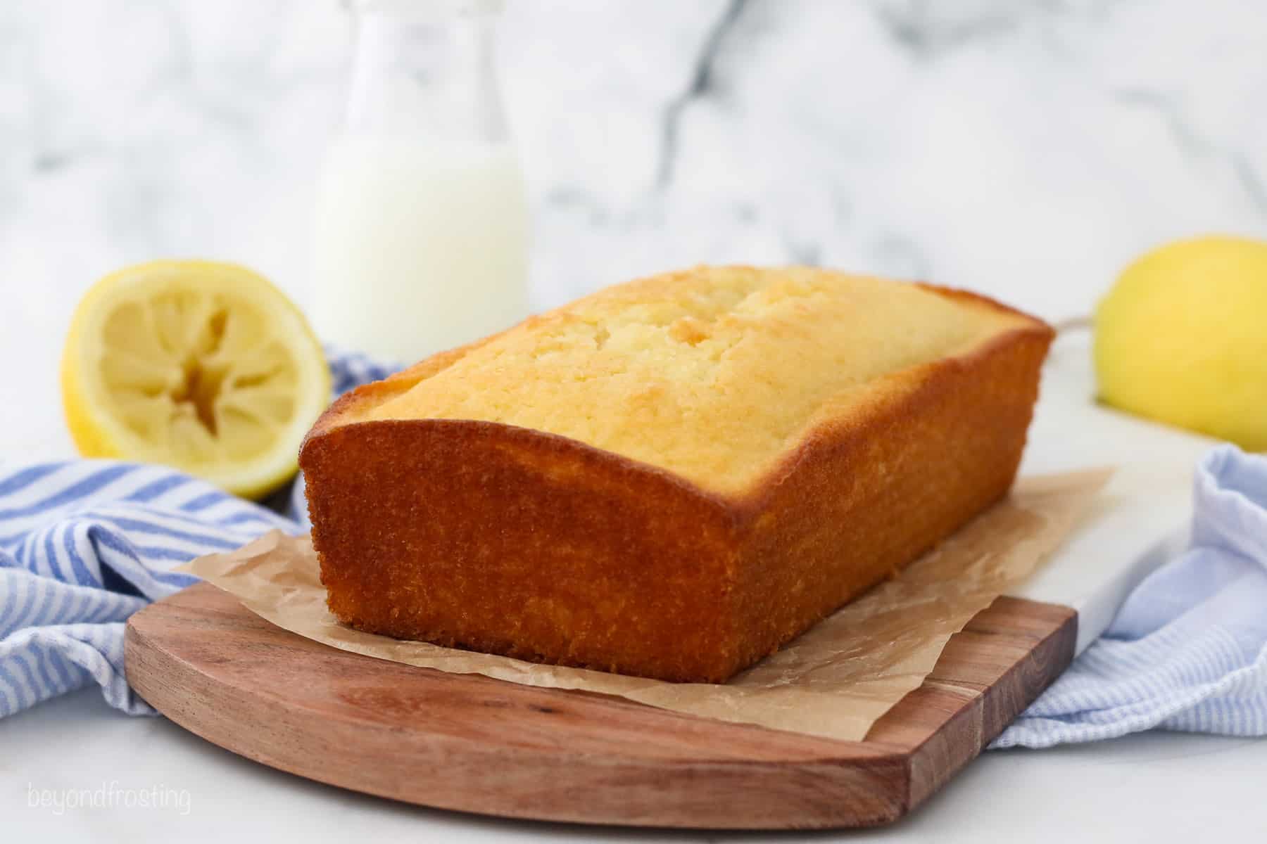 A baked lemon loaf on a wooden cutting board lined with parchment paper, with lemon halves in the background.