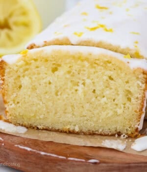 Glazed lemon bread with a slice cut from the end on a wooden cutting board lined with parchment paper, with a lemon half in the background.
