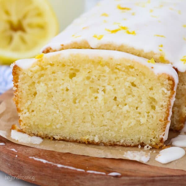 Glazed lemon bread with a slice cut from the end on a wooden cutting board lined with parchment paper, with a lemon half in the background.