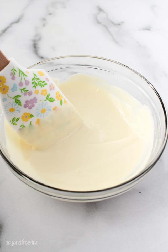 a floral spatula stirring melted white chocolate in a glass bowl