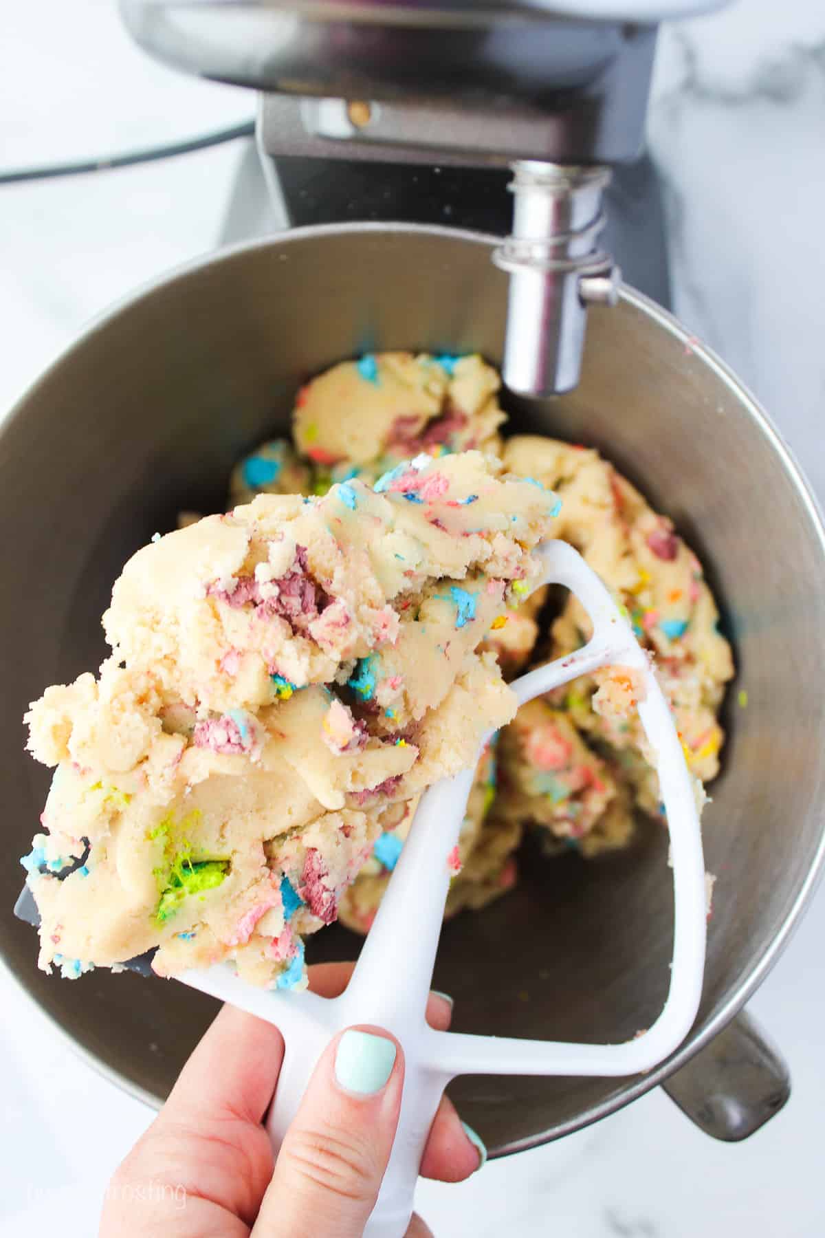 A hand holding a stand mixer attachment covered with Lucky Charms cookie dough over dough in a metal mixing bowl.