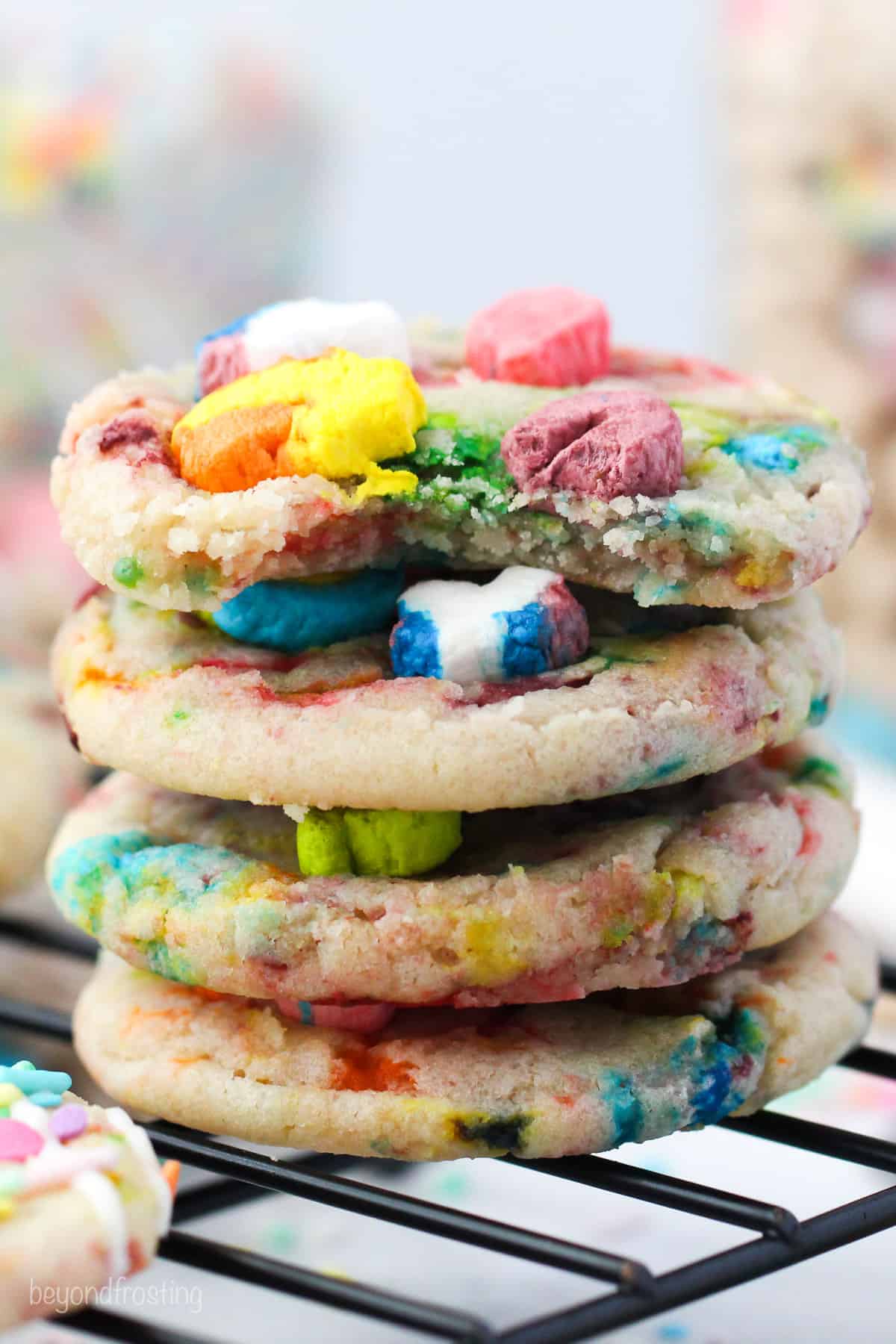 Four Lucky Charms cookies stacked on a wire rack, with a bite missing from the top cookie.