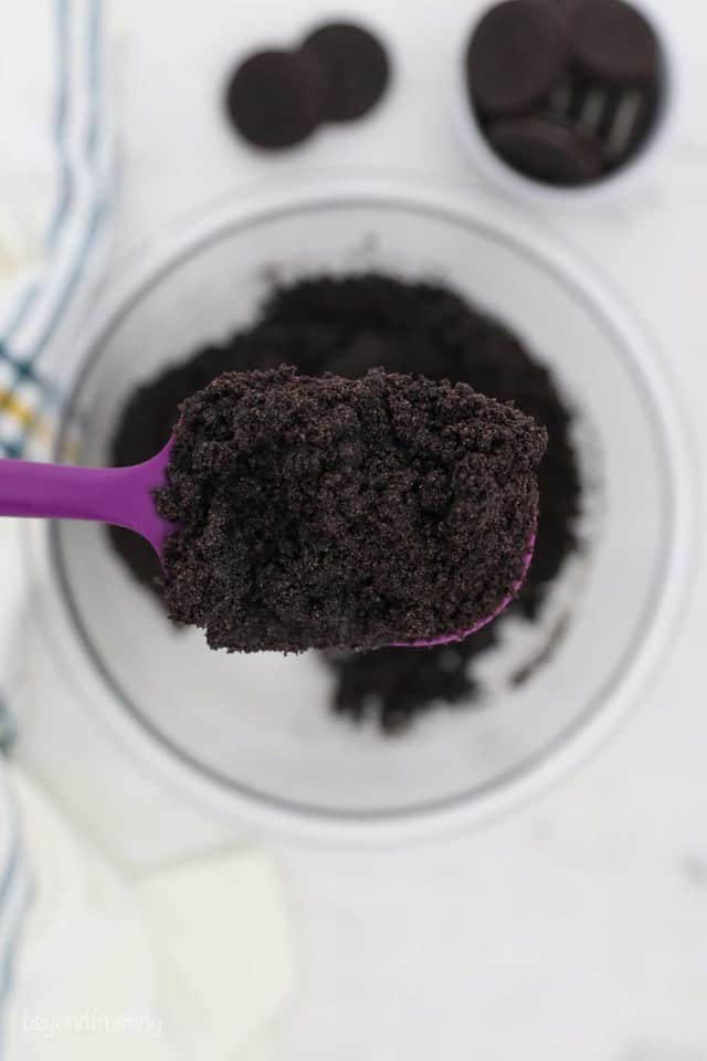 A Purple Spatula Holding Oreo Cookie Crumbs Over a Glass Mixing Bowl
