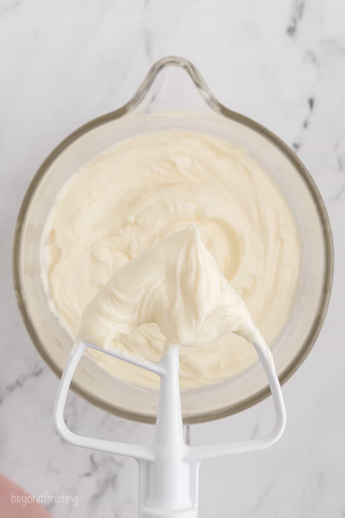 A stand mixer attachment covered with cream cheese frosting held over frosting in a mixing bowl.