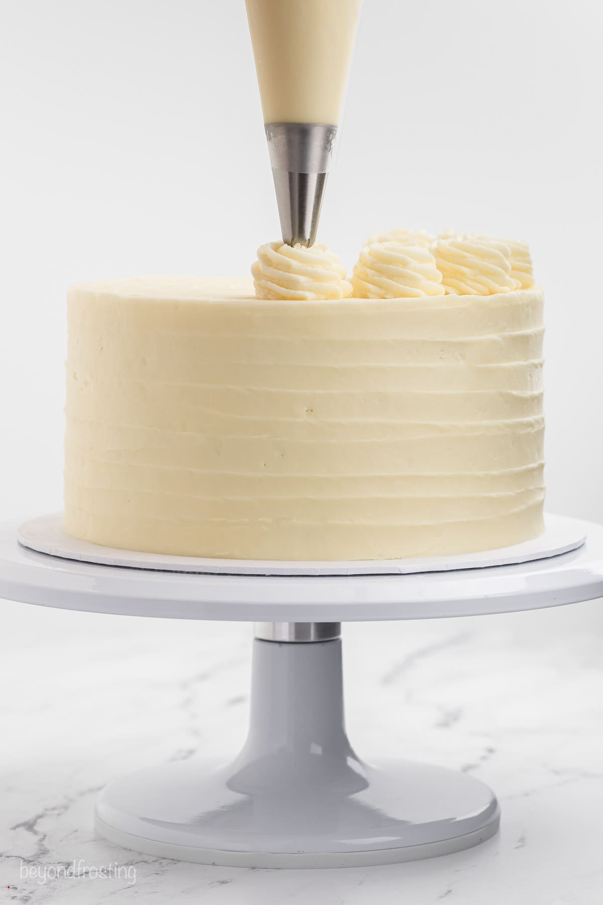 A piping bag pipes swirls of cream cheese frosting over a frosted red velvet layer cake on a cake stand.