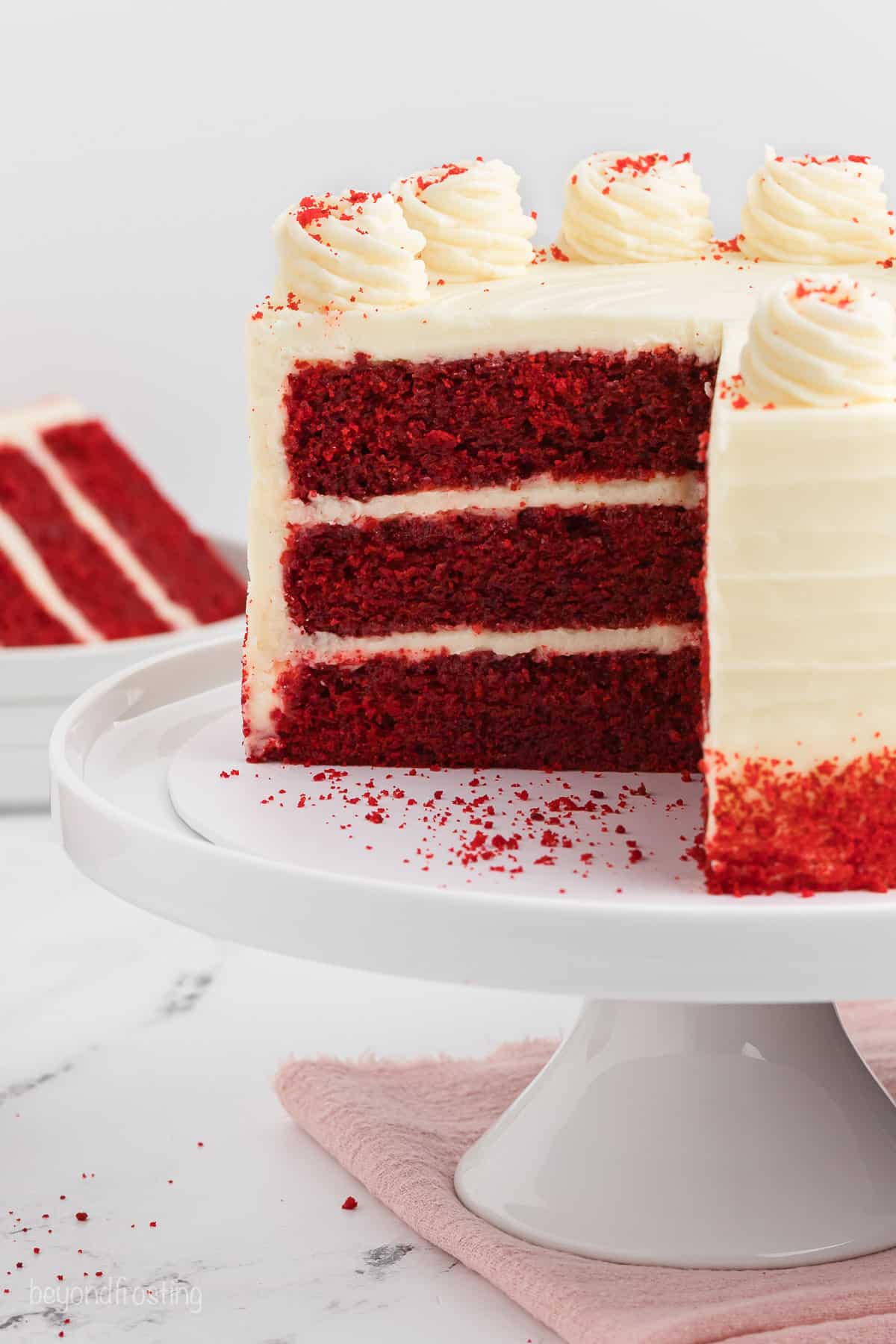 Frosted red velvet layer cake on a cake stand, with a large slice missing.