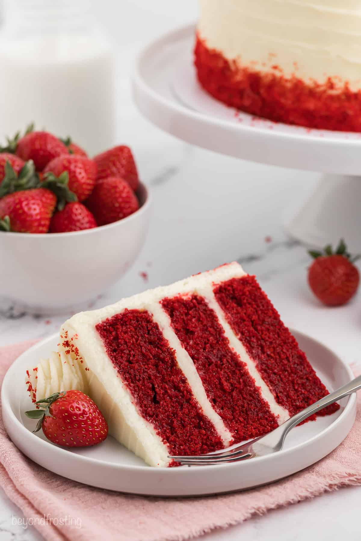 A slice of three-layer frosted red velvet cake on a plate next to a fork, with a bowl of strawberries and the rest of the cake in the background.