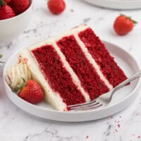 A slice of three-layer frosted red velvet cake on a plate next to a fork.