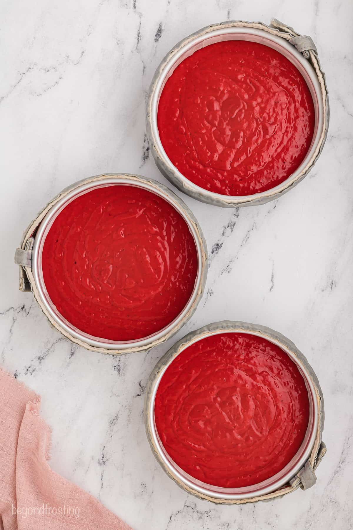 Overhead view of three round cake pans filled with red velvet cake batter.