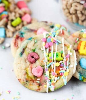 A Lucky Charms cookie drizzled with icing propped up against a stack of cookies on a white countertop, with Lucky Charms cereal in the background.