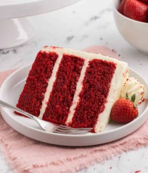 A slice of three-layer frosted red velvet cake on a plate next to a fork.