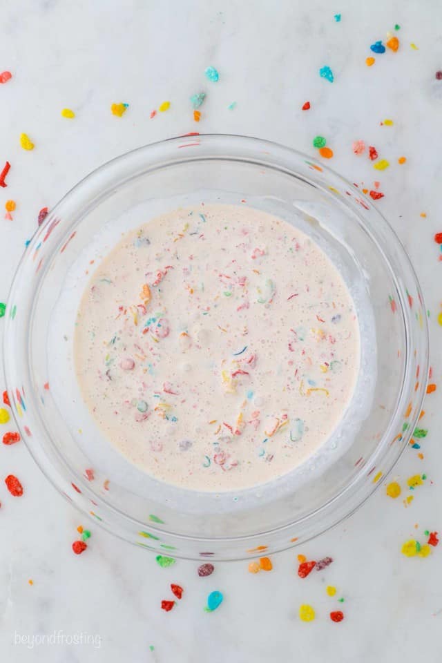 Bird's eye view of a glass mixing bowl with heavy cream and fruity pebble cereal