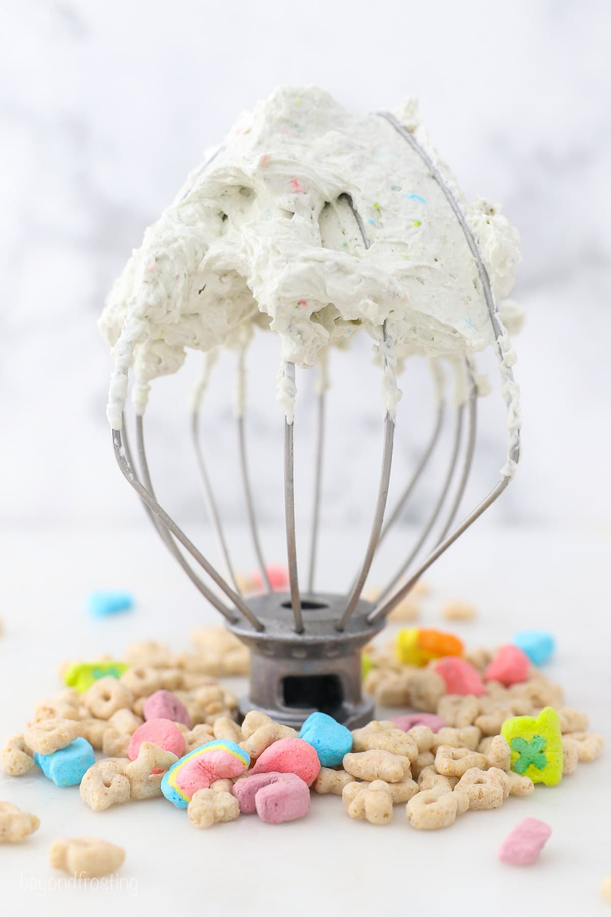 A stand mixer attachment topped with cereal milk whipped cream standing upright on a countertop, surrounded by Lucky Charms cereal.