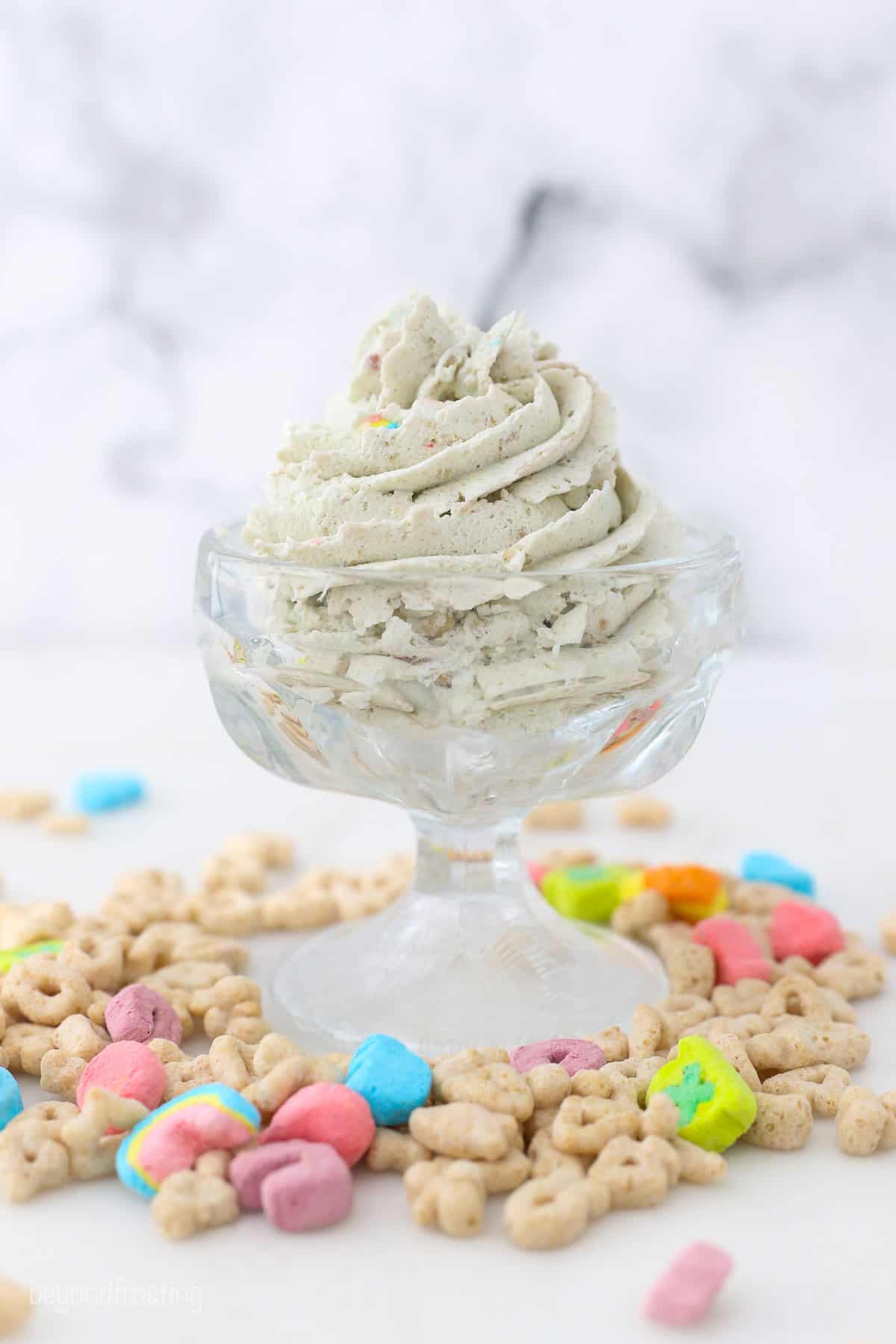 A piped swirl of cereal milk whipped cream in a glass bowl on a countertop, surrounded by Lucky Charms cereal.