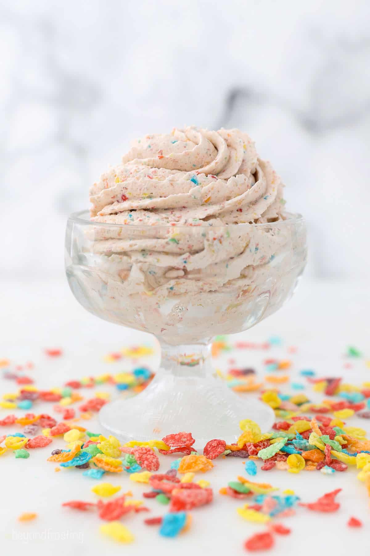 A piped swirl of cereal milk whipped cream in a glass bowl on a countertop, surrounded by Fruity Pebbles cereal.