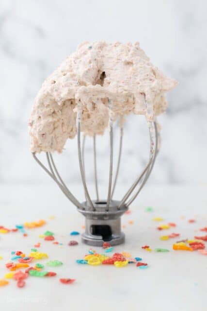 a wire whisk with whipped cream surrounded by Fruity Pebbles Cereal
