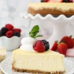 A slice of cheesecake topped with whipped cream and berries. In the back is the full cheesecake.